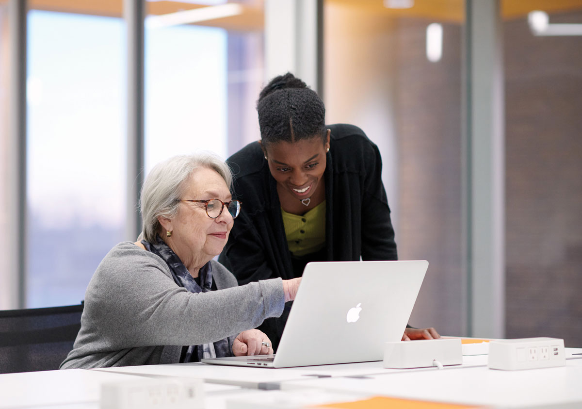 A young woman leans over a senior woman's shoulder to look at a laptop