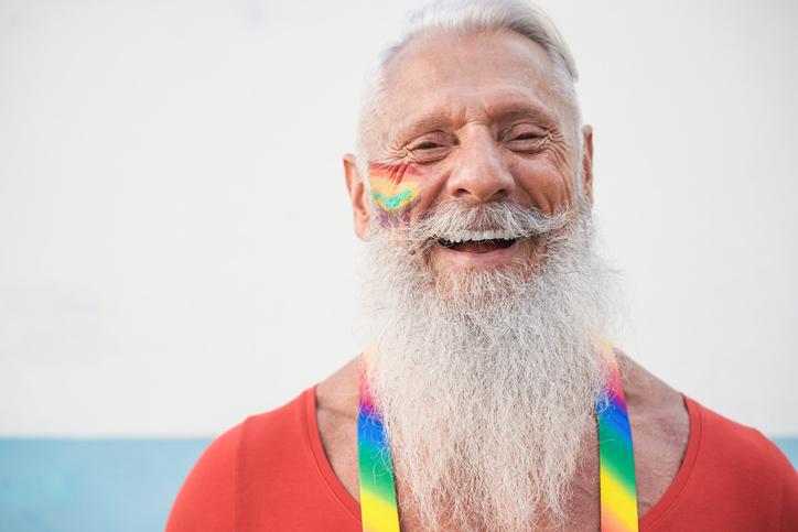 Smiling elder gay man with gray beard has a rainbow on his cheek and a rainbow tie draped around his shoulders